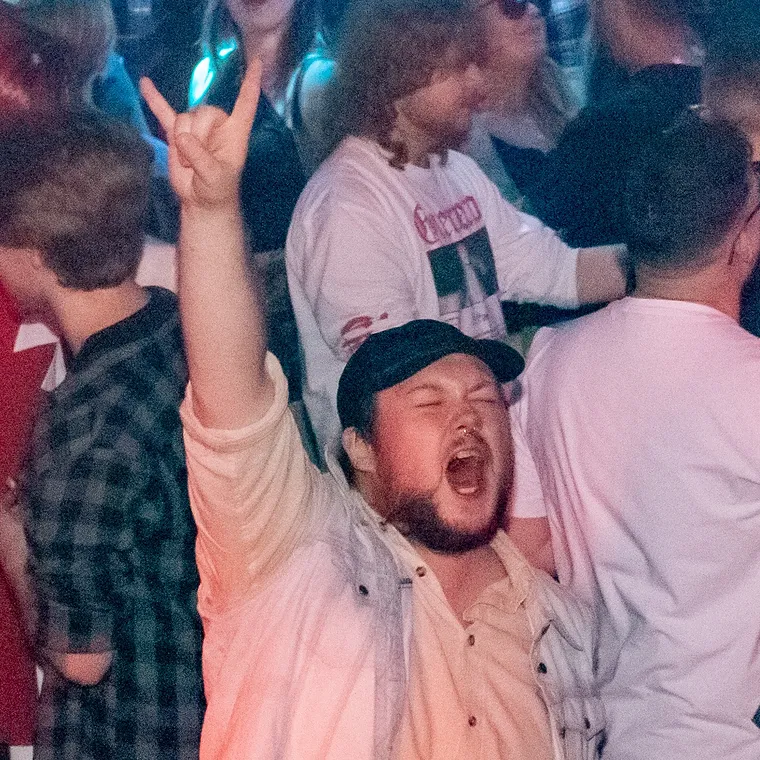 A man in a cap mid scream with his horns up, the crowd going around him